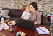أم عاملة تحمل طفلتها وتعمل على اللابتوب وسط مهام المنزل اليومية - صورة تعبيرية
